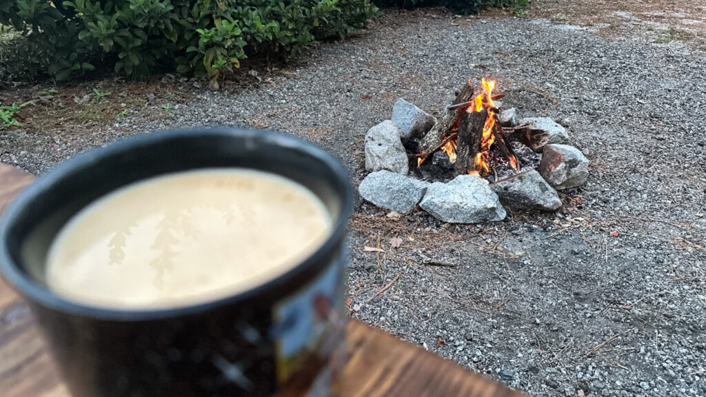 “Close-up of a warm coffee cup on a wooden table with a cozy campfire burning in a stone fire ring in the background. Surrounded by greenery, this peaceful scene captures the simple joys of camping: a hot drink and a morning fire in nature.”